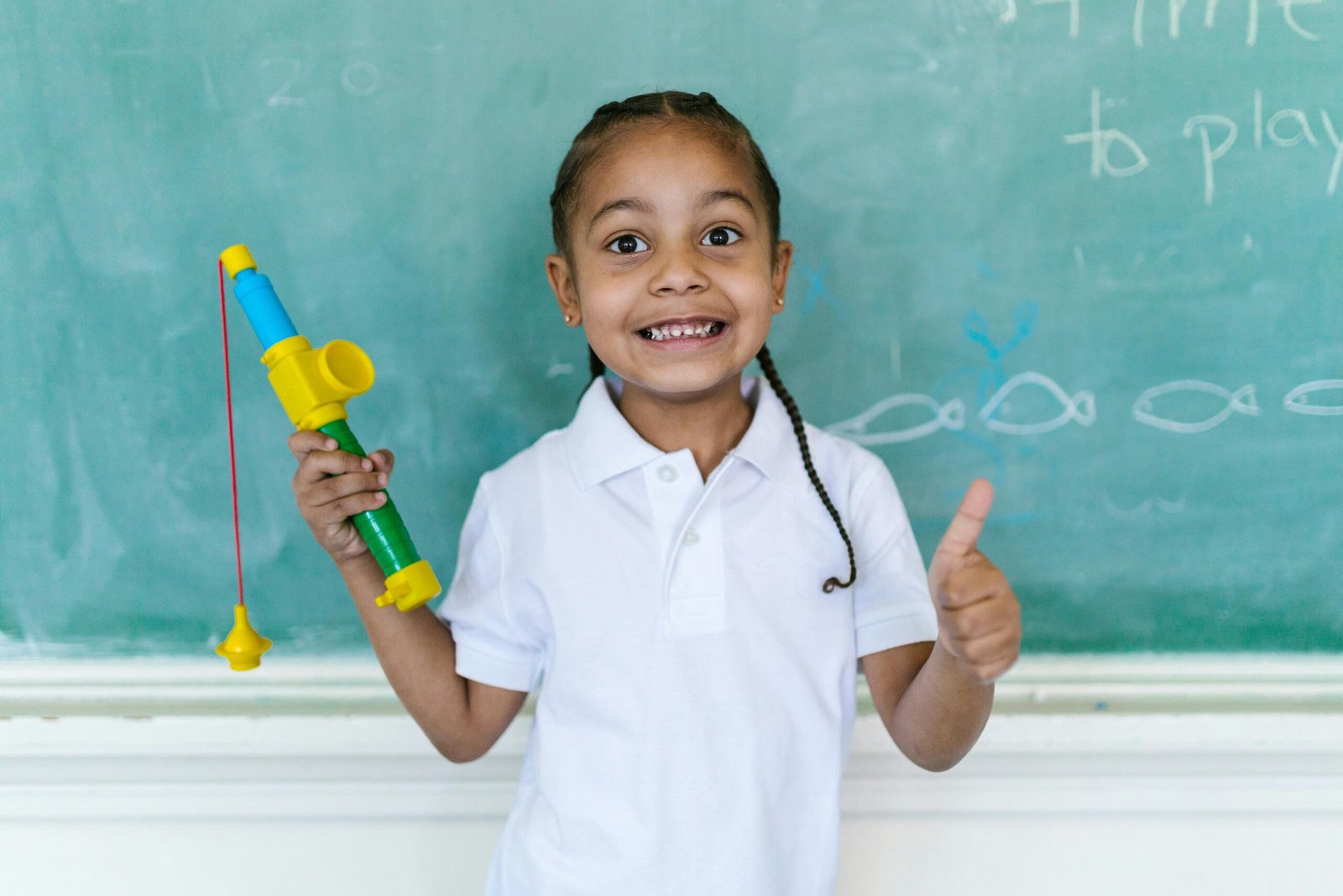 Smiling child in a white polo shirt giving a thumbs up in front of a chalkboard, holding a toy fishing rod.